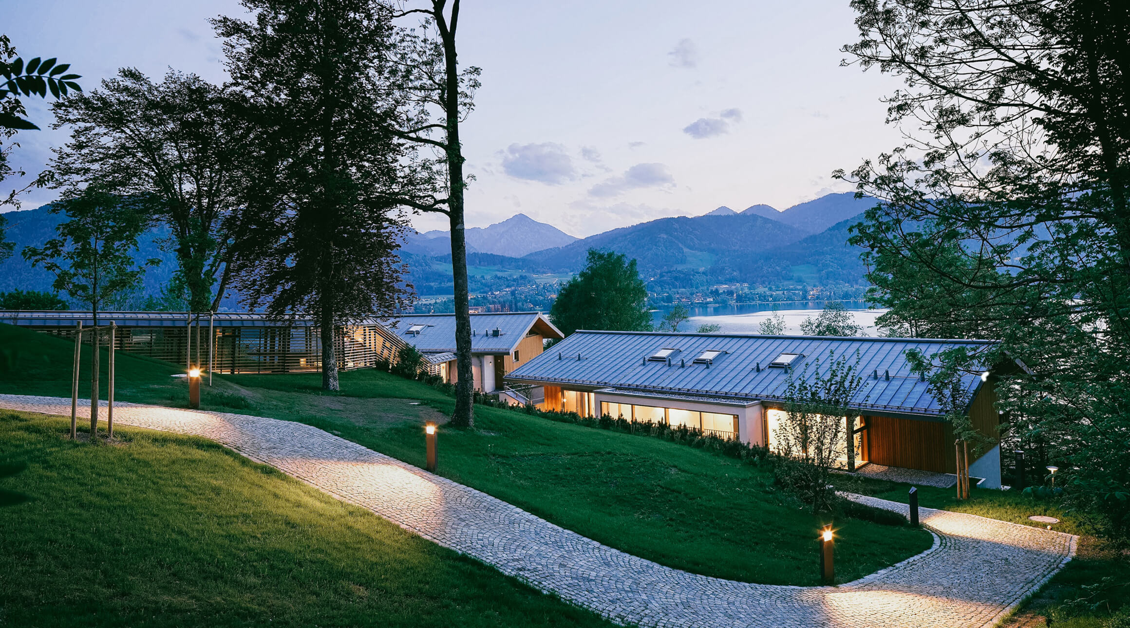 Blick von außen auf die Alpenchalets direkt am Tegernsee mit Blick auf die umliegenden Berge in der Abenddämmerung.