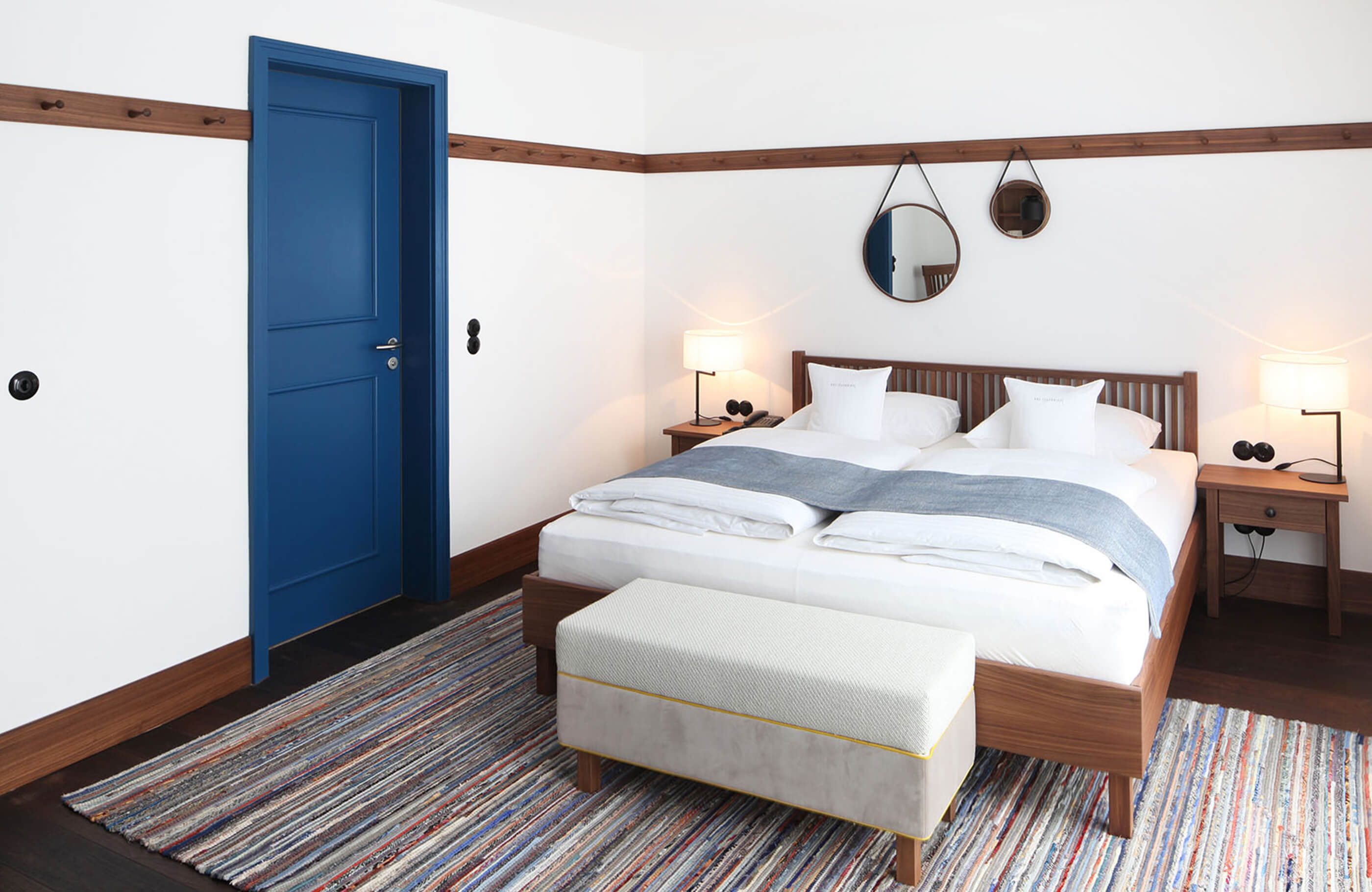 Aufnahme des puristischen Doppelzimmers mit einem großen Doppelbett, rechts und links davon jeweils ein Nachtkästchen mit Tischleuchte und einer Truhensitzbank vor dem Bett.