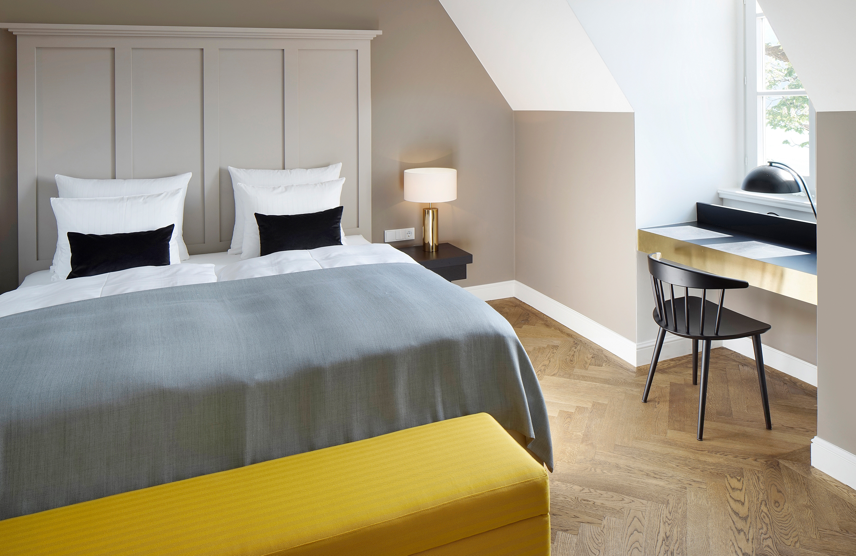 Blick auf ein Bett mit einer gelben gepolsterten Sitzbank am Ende und einem kleinen Schreibtisch