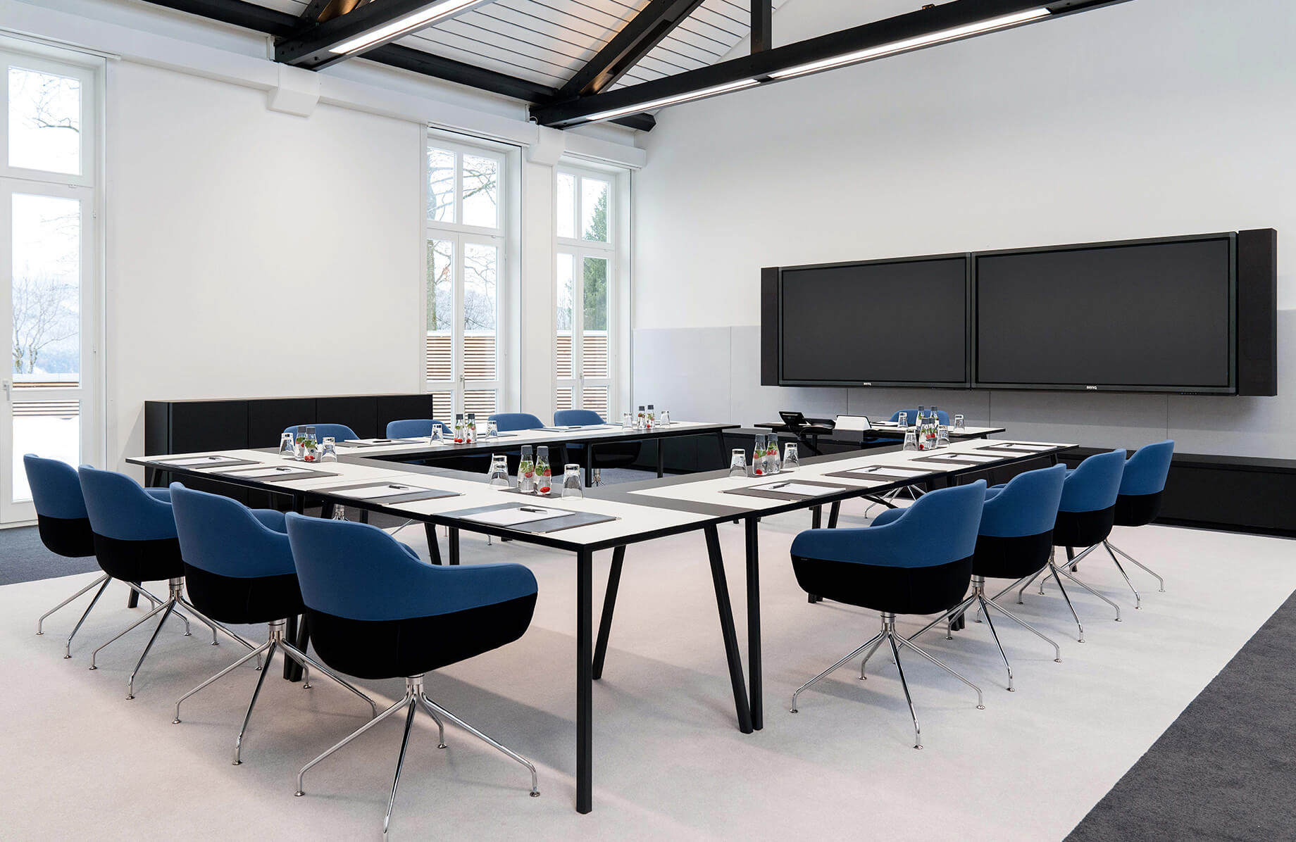 Aufnahme eines Tagungsraumes im Haus Wallberg, in dem mehrere Tische und blau-schwarze Stühle in U-Form vor einem schwarzen Bildschirm aufgebaut sind.