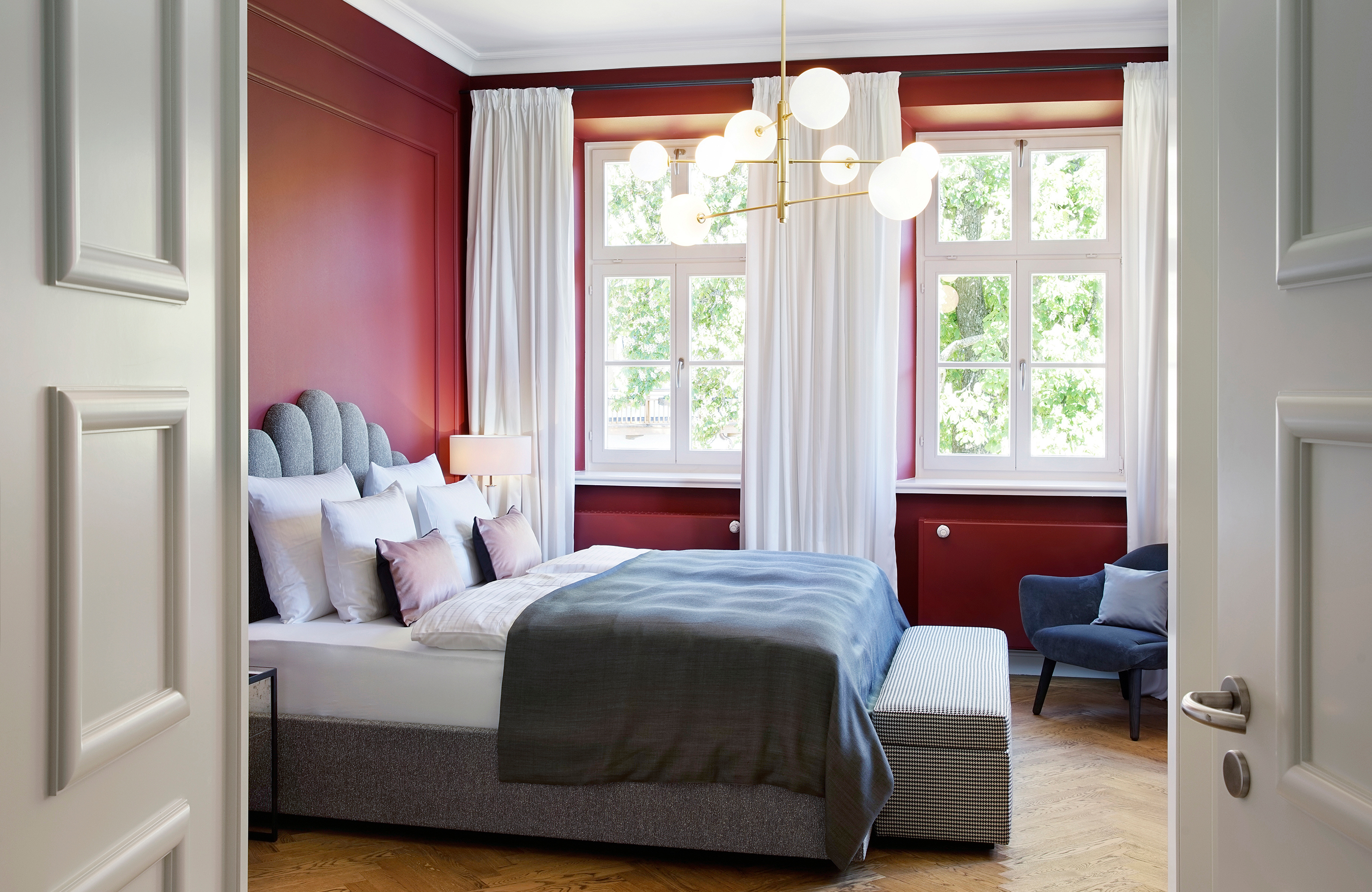 Blick in den Schlafbereich der Superior Suite im Hotel Das Tegernsee mit einem großen, hellgrauen Bett im Zentrum des Raums, einer Truhensitzbank vor dem Bett und einem grauen Sessel vor einer hellen Fensterfront mit Blick nach draußen.