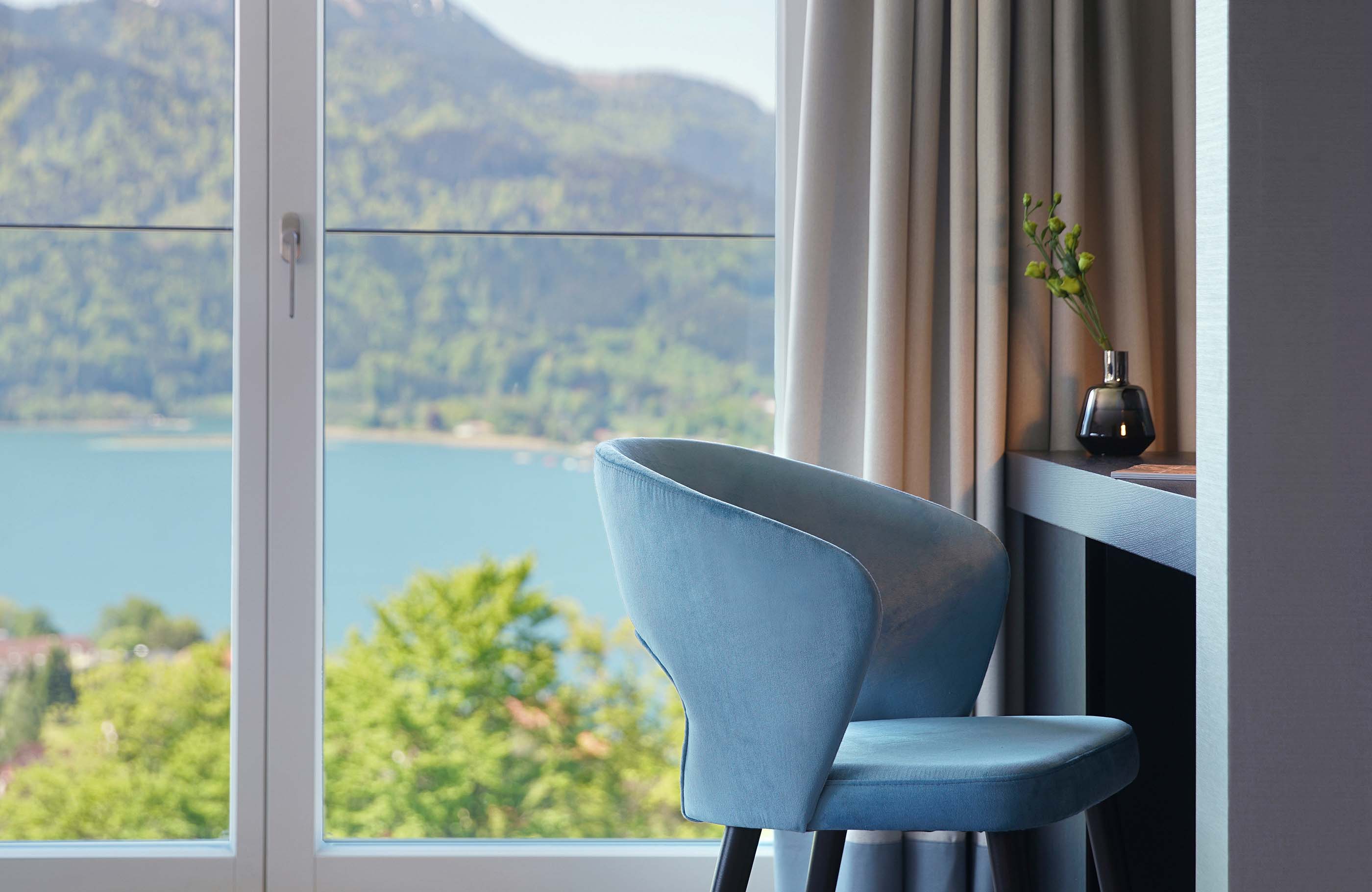 Detailaufnahme des blauen Schreibtischstuhls vor einem Fenster mit Blick auf den Tegernsee