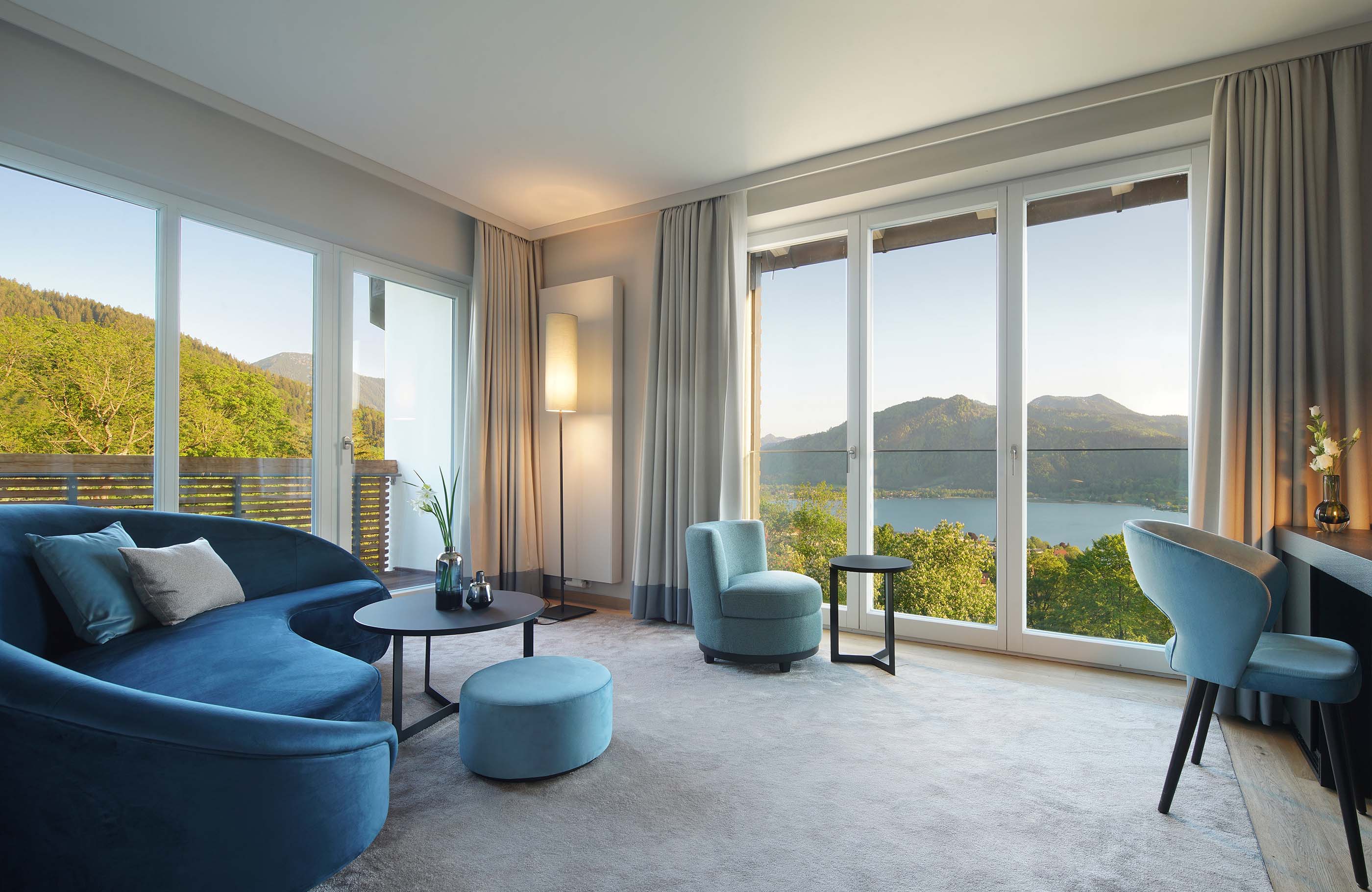 Aufnahme des Wohnzimmers mit blauen Möbeln und zwei großen Fensterfronten mit Blick über den Tegernsee