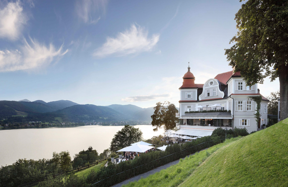 Die Außenansicht des Hotels Tegernsee und die Terrasse mit Gästen, die am Tegernsee frühstücken