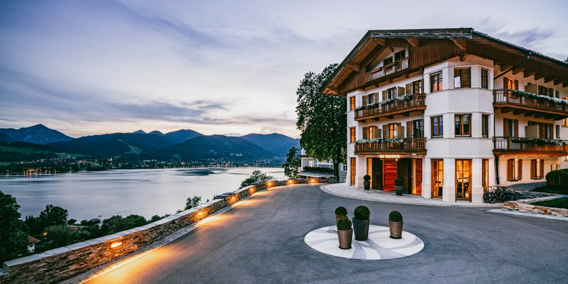 Aufnahme von der Hotelauffahrt und dem Haus Tegernsee von außen mit Blick auf den Tegernsee und die umliegenden Berge während der Abenddämmerung.